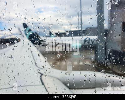 View from inside an airplane cabin looking out through a rain-covered window onto the airport tarmac. Water droplets of various sizes cover the glass Stock Photo