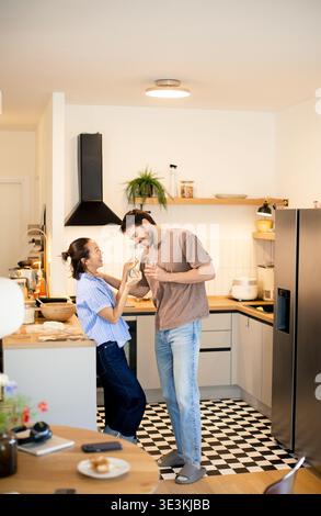 Two people are having fun in a kitchen. They are cooking together and laughing. The kitchen has modern features and light colors. Food is seen on the Stock Photo