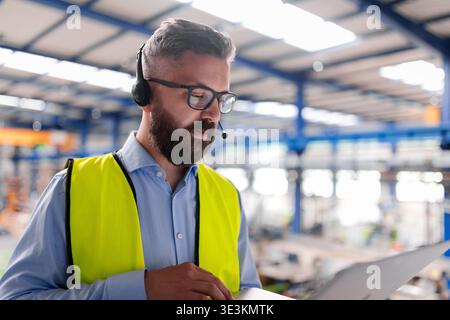Quality manager with headset and laptop standing in industrial factory. Stock Photo