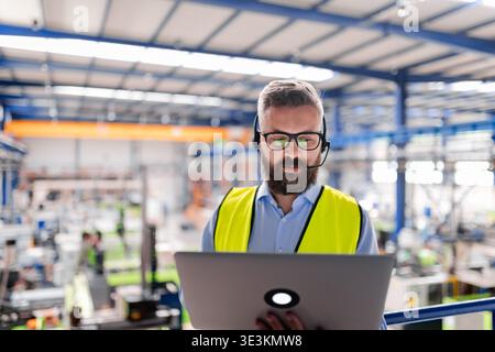 Quality manager with headset and laptop standing in industrial factory. Stock Photo