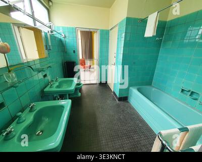 A vintage bathroom interior featuring turquoise ceramic wall tiles and matching green fixtures. The room contains two wall-mounted sinks with chrome Stock Photo