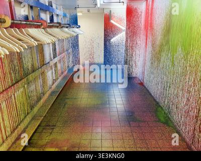 A narrow interior corridor with walls, floor, and ceiling covered in vibrant, dripping paint splatters in rainbow colors including red, blue, green, Stock Photo