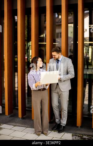 Two people stand outside a modern building in urban setting. The man and woman discuss a document while casually engaging in conversation. It is dayti Stock Photo