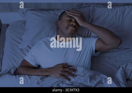 Black man lying in bed at night holding head, suffering from headache Stock Photo