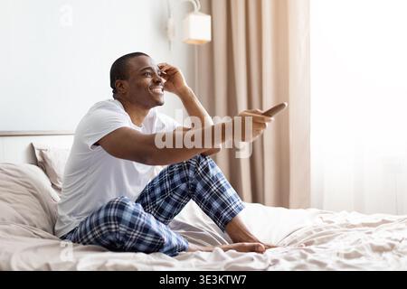 Young black man sitting on bed smiling while holding remote control Stock Photo
