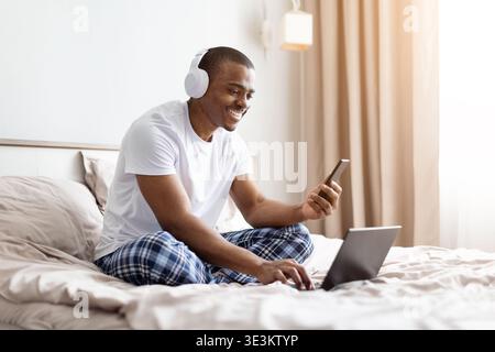 Young black man sitting on bed using smartphone and laptop while smiling Stock Photo
