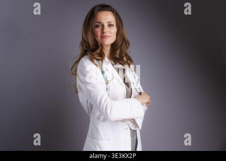 A medium-shot portrait of a focused middle-aged female medical specialist in a white lab coat and stethoscope, posing with crossed arms against a dark Stock Photo