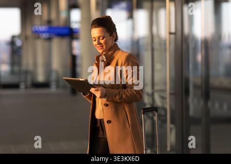 A focused woman in a tan coat stands in a modern transit hub, using her tablet to check travel notifications and boarding information before her train Stock Photo