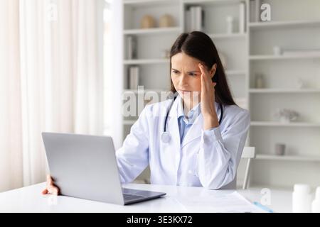 Stressed female doctor working on laptop in clinic office, touching her temple Stock Photo