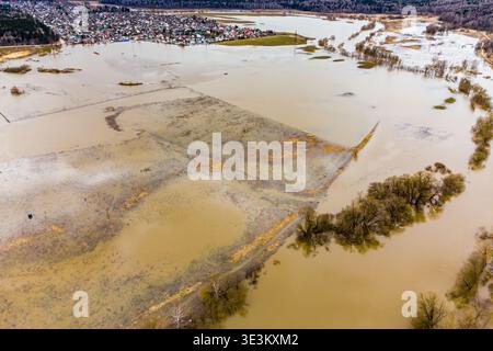 Panoramic drone view captures a mighty spring deluge, muddy river bursting its banks, swallowing fields. Remote settlement peeks through the haze, a t Stock Photo