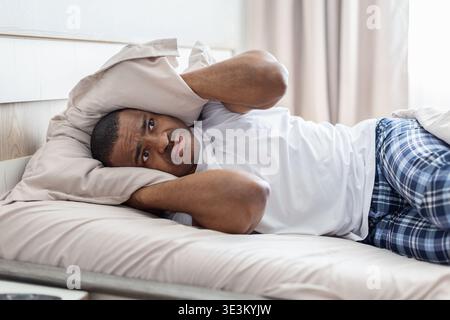 Young african american man lying in bed covering ears with pillow Stock Photo