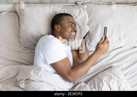 Black man lying in bed smiling while using smartphone, enjoying digital content Stock Photo