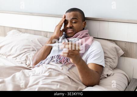 Young black man lying in bed checking temperature with thermometer Stock Photo
