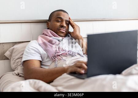 Young african american man lying in bed working on laptop while sick Stock Photo