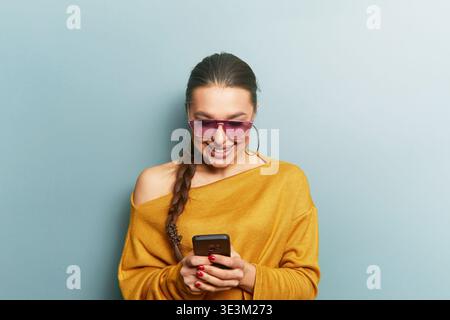 Digital lifestyle concept. Young woman holding smartphone. Smiling while texting. Casual outfit with sunglasses. Portrait focusing on face and interac Stock Photo