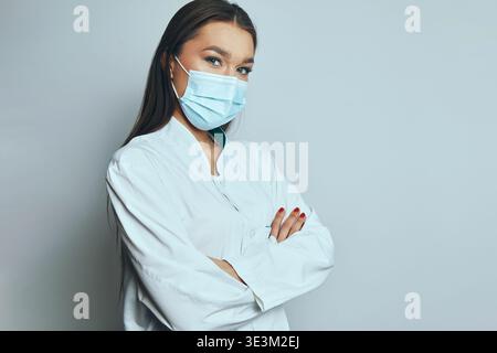 A healthcare worker stands with arms crossed, wearing a mask and looking directly at the viewer. The background is plain and light. The worker shows a Stock Photo