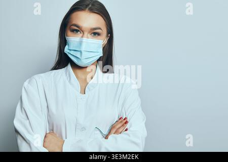 A health worker stands with arms crossed wearing a mask in a clinical space. The setting has a simple background. The worker appears focused and serio Stock Photo