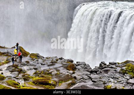 Water pours down Dettifoss waterfall in Iceland as visitors walk along rocks near the edge. It is a cloudy day with mist in the air. Stock Photo