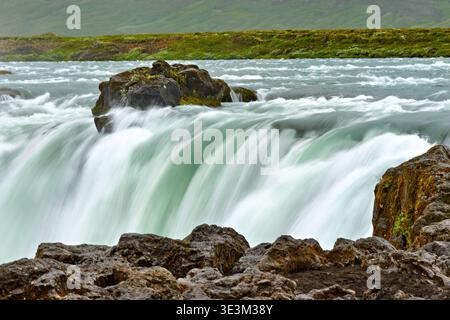 Water rushes over rocks at Godafoss waterfall in Iceland. The scenery shows strong water flow and green land in the background. Stock Photo
