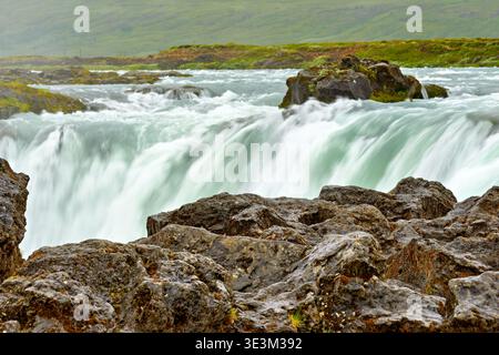 Water flows over the rocks at Godafoss waterfall in Iceland. People gather nearby to enjoy the sight of the water cascading down. Stock Photo
