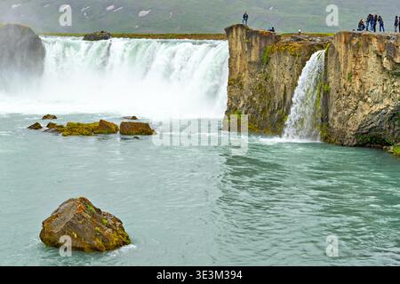 Water flows over the edge of Godafoss Waterfall in Iceland. People explore the area, enjoying the view and taking photos of the waterfall. Stock Photo