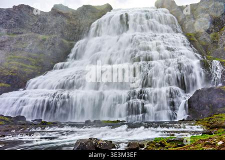 People watch water flow down Dynjandi Waterfall in Iceland. The waterfall is part of the Westfjords region and has several tiers. Stock Photo
