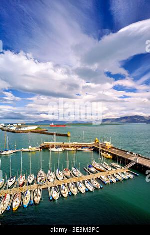 Sailboats are parked at Reykjavik harbor. The clouds create a dramatic sky above the water. Nearby buildings and mountains add to the view. Stock Photo