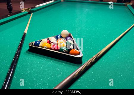 Balls sit in triangle on green felt of billiard table with cues placed nearby. Good lighting highlights various colors and textures, showing a game of Stock Photo