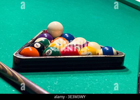 Billiard table shows balls arranged in a triangle with cue stick nearby. Lighting highlights the green felt and wooden edges, setting up for game and Stock Photo
