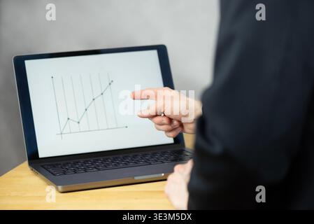 A man is standing at a desk and looking on a laptop screenwhich displays a line chart. The focus is on the laptop on the desk and the persons hands. Stock Photo