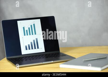 A laptop is put on a desk. The screen shows two line graphs displaying some company’s statistics. A notebook and a pen are lying next to the laptop. Stock Photo