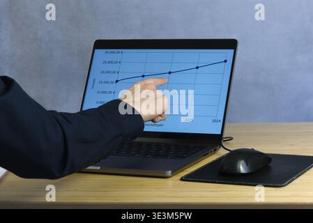 A businessman is standing at a table. He is pointing to a chart on a laptop screen showing the company's development with a positive trend curve. Stock Photo