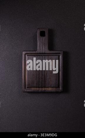 Empty rectangular serving board made of dark brown wood resting on a textured black surface, ready for food preparation, cutting, or stylish presentat Stock Photo