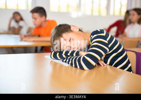 Bored tween schoolboy sleeping at desk in classroom during lesson Stock Photo