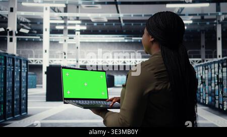 Engineer at work using green screen laptop in data center to check hardware for system stability. Worker using mockup notebook to oversee server farm and configure infrastructure, camera A Stock Photo