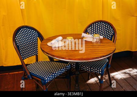 Two chairs and a round table set for dining in front of a bright yellow backdrop at a cafe during daytime on a sunny day Stock Photo
