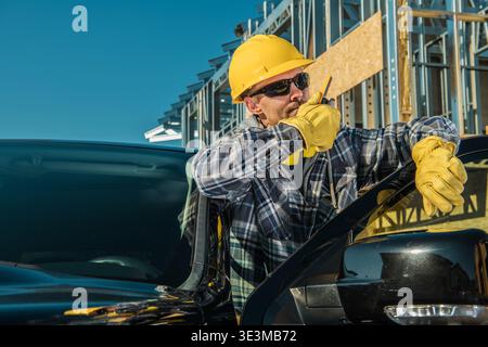 A worker wearing a yellow hard hat stands by a black truck. He holds a radio and looks at plans. Stock Photo