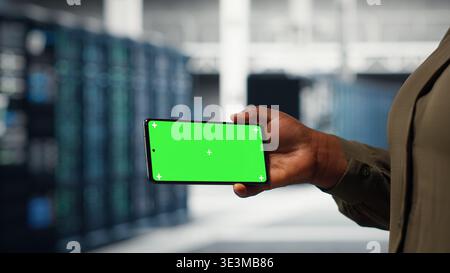 Software developer using app on mockup phone to check server hub energy consumption. African american woman using green screen smartphone in data center to do gear checkup, camera B Stock Photo