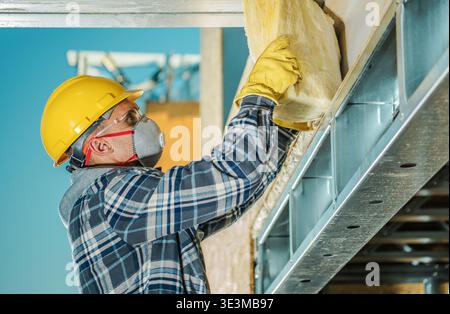 A worker wearing a hard hat and mask installs insulation in a building. He uses gloves for safety. Stock Photo
