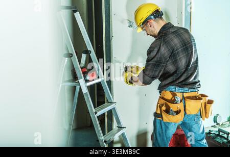 A worker is fixing drywall at a construction site using tools. A ladder stands nearby. Stock Photo