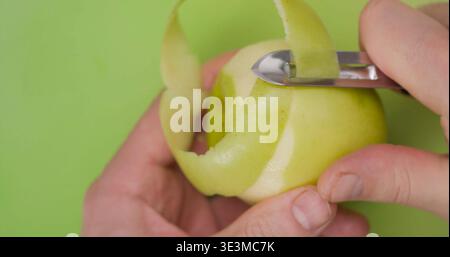 Hands peel a green apple with a knife on a light green cutting board in a kitchen setting during the day Stock Photo
