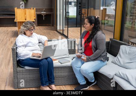 Two women relaxing on outdoor sofa, having a conversation and working on a laptop Stock Photo