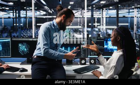 Manager supervising woman in data center coding with generative AI assistance. Supervisor oversees work by african american IT expert leveraging artificial intelligence large language model, camera A Stock Photo
