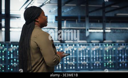 Data center programmer uses tablet to develop disaster recovery plans, minimizing downtime. IT professional walking in server farm, enhancing data protection protocols using device, camera B Stock Photo