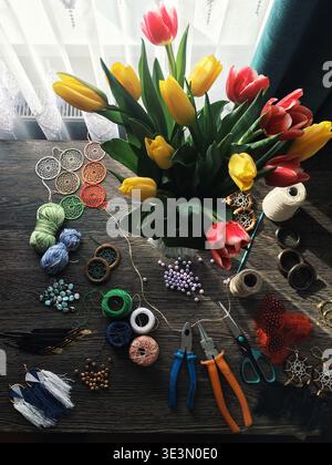 A bouquet of tulips in red and yellow sits on a wooden table Various crafting supplies like threads beads and tools are spread out around the flowers Stock Photo