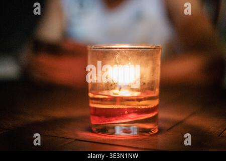 Warm candle glows softly in a glass. Candlelight casts gentle, golden reflections. Glass rests on dark, textured wooden surface. Soft bokeh background Stock Photo