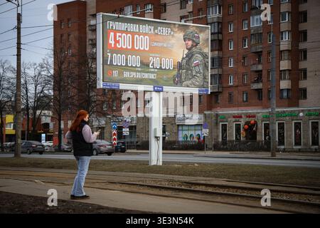 A woman stands in front of an information billboard advertising contract service in the Russian Armed Forces, installed on one of the city streets. A large-scale information campaign to recruit citizens for contract military service continues on the streets of St. Petersburg. Billboards and billboards throughout the city display up-to-date information on recruitment for various units of the Russian Armed Forces. Outdoor advertising places particular emphasis on financial terms: billboards list the one-time payment amounts, which will reach 4,500,000 rubles in March 2026. Campaign materials als Stock Photo