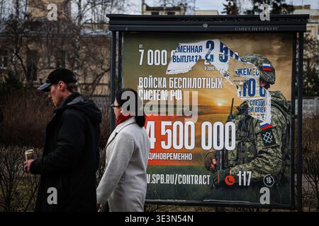 A man and a woman walk past an information billboard advertising contract service in the Russian Armed Forces, installed on one of the city streets. A large-scale information campaign to recruit citizens for contract military service continues on the streets of St. Petersburg. Billboards and billboards throughout the city display up-to-date information on recruitment for various units of the Russian Armed Forces. Outdoor advertising places particular emphasis on financial terms: billboards list the one-time payment amounts, which will reach 4,500,000 rubles in March 2026. Campaign materials al Stock Photo