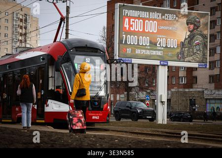 Residents stand at a tram stop in front of an information billboard advertising contract service in the Russian Armed Forces, which is installed on one of the city streets. A large-scale information campaign to recruit citizens for contract military service continues on the streets of St. Petersburg. Billboards and billboards throughout the city display up-to-date information on recruitment for various units of the Russian Armed Forces. Outdoor advertising places particular emphasis on financial terms: billboards list the one-time payment amounts, which will reach 4,500,000 rubles in March 202 Stock Photo