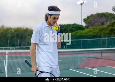 Asian man standing on hard tennis court holding racket and yellow ball, wearing white headband. Athlete, outdoor, hardcourt, focus, solitude, fence, f Stock Photo
